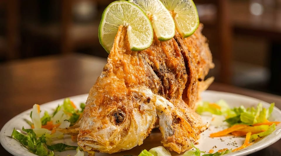 Whole fried fish, garnished with lime slices and a side salad, on a white plate.